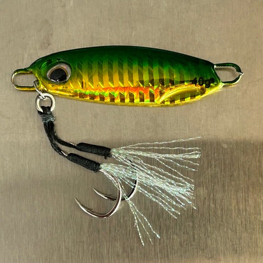 A metallic yellow and green fishing lure with a black treble hook and white skirt, placed on a light wood plank surface.