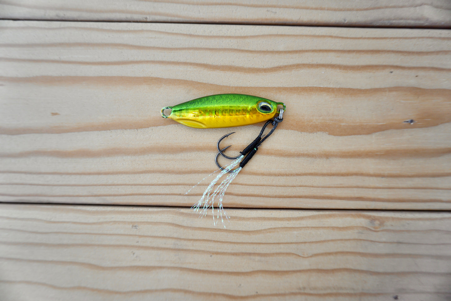 A metallic yellow and green fishing lure with a black treble hook and white skirt, placed on a light wood plank surface.